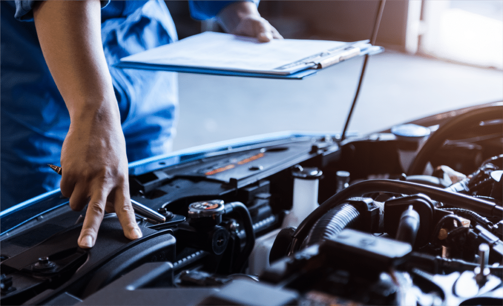 Description: Mechanic in blue coveralls inspecting a car engine with a clipboard and pen in hand at Horeb Auto Repair.